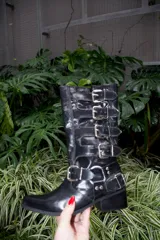 Bota de caña alta estilo biker color rojo cereza, confeccionada en cuero vacuno. Presenta múltiples tiras horizontales con hebillas metálicas a lo largo de la caña y una tira adicional con hebilla y herrajes metálicos en la zona del tobillo. Cuenta con taco cuadrado de madera y punta ligeramente afinada.