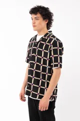 Camisa de manga corta con calce relajado y cuello tipo sastre, confeccionada en rayón. Presenta un diseño de cuadrícula beige sobre fondo negro con detalles en azul y rojo en las intersecciones.