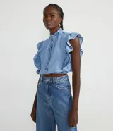 Camisa cropped de jeans celeste, sin mangas, con cuello alto, cierre frontal con botones, volantes en cuello y laterales, y elástico en la cintura.