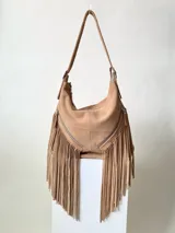 Bolso tipo hobo color tostado, hecho de cuero con flecos largos en el frente y dos bolsillos con cierre. Tiene una manija regulable con hebillas metálicas.