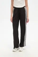 Pantalón de algodón negro, holgado, de calce medio, con cintura elástica y cordón ajustable. Presenta tres rayas blancas verticales en los laterales.