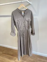 Vestido camisero largo color gris topo de pana fina con cuello mao, cierre frontal con botones, mangas largas con puños abotonados, bolsillos de parche en el pecho, cintura elastizada con cordón ajustable y bolsillos laterales.