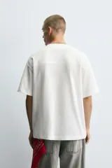 Camiseta blanca de corte holgado con cuello en V y mangas cortas. Presenta un diseño de malla con diferentes texturas combinadas a tono y estampado frontal con la inscripción "Legacy Crew 95 Elite Mode".