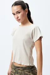 Remera color beige de punto acanalado con silueta relajada y manga corta raglán. El detalle de ruedo con terminación ondulada le aporta un aire fresco y juvenil.