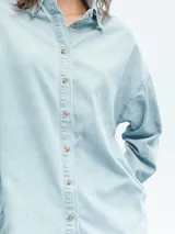 Camisa celeste de denim con botones marrones, corte holgado y mangas largas.
