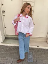Camisa blanca de corte holgado con cuello camisero y mangas largas con puños elásticos. Presenta bolsillo de parche en el pecho y detalles de ribete rojo en cuello, puños y bolsillo.