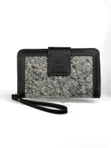 Billetera tipo clutch o cartera de mano, con cuerpo principal de cuero negro texturizado y panel central de pelo (efecto vaca) en tonos blanco y negro. Posee cierre de solapa con broche y correa de muñeca desmontable en cuero negro.