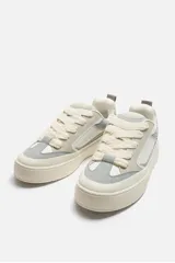 Zapatilla deportiva tipo skate con parte superior fabricada en combinación de piezas y materiales. Detalle de cordones maxi y suela con volumen. Color blanco, gris y crema.
