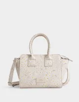 Cartera mini city de cuerina color beige con estampado de flores blancas y amarillas. Tiene correa desmontable de cuerina y doble asa corta de mano. El acceso es mediante cierre superior y tiene un tag de cuerina con la inscripción "Enjoy the now".
