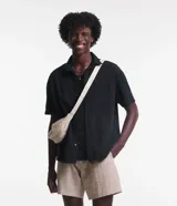 Camisa negra de sarga, corte boxy, con cuello inglés, manga corta, abotonadura frontal y bolsillo en el pecho.