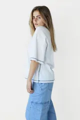 Remera blanca de corte holgado con cuello redondo y mangas cortas. Presenta un pequeño estampado de una flor amarilla en el centro del pecho y detalles de costura en azul en cuello, mangas y ruedo.