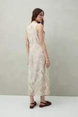 Vestido largo sin mangas con diseño cruzado y lazo lateral. Confeccionado en mezcla de lino y viscosa, presenta un estampado sutil de hojas en tonos marrones sobre un fondo beige y una abertura frontal que aporta movimiento.