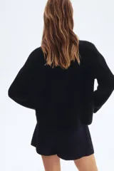 Cárdigan corto negro de punto acanalado, con cuello redondo, manga larga y botones metálicos dorados en el frente. Posee bolsillos de parche con botón en la parte inferior delantera.