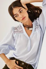 Camisa de algodón color celeste, con escote en V, cierre parcial de botones y detalles de encaje en los hombros. Mangas largas abullonadas con puños elásticos.