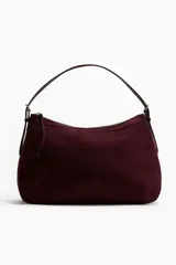 Bolso de hombro color borgoña, con textura que simula gamuza o ante, de forma curva y cierre superior con cremallera. Posee una correa de hombro delgada de cuero marrón oscuro y un tirador de cremallera con borla a juego.