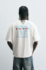 Camiseta color crudo de corte relajado, cuello redondo y manga corta. Estampado frontal con texto "Flies to Los Angeles CA" y diseño de bandera a cuadros.
