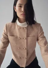 Chaqueta corta de tweed color rosa pálido con doble botonadura dorada, cuello redondo y bolsillos de parche en el pecho.