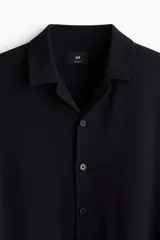 Camisa negra de tejido suave con cuello abierto, tapeta lisa y canesú en la espalda. Modelo de manga larga con cierre ajustable de botones en los puños y tapeta con botón. Bajo recto y corte estándar.
