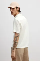 Camisa blanca de punto con textura acanalada, corte regular, cuello inglés, tapeta lisa, canesú en la espalda y bajo recto con aberturas laterales.