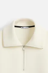 Sudadera color crudo de tejido técnico con cuello alto y cierre de cremallera. Tiene manga larga, corte relaxed fit, bolsillo tipo canguro y rib en puños y bajo con rayas negras.