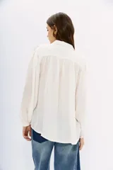 Camisa blanca de manga larga confeccionada en viscosa con acabado sandwash, de textura suave y aterciopelada. Cuello camisero con botón en forma de corazón y frunces en el canesú delantero y la espalda.