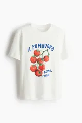 Remera blanca de corte holgado con estampado de tomates y texto en azul. Tiene cuello redondo ribeteado acanalado y hombros caídos.