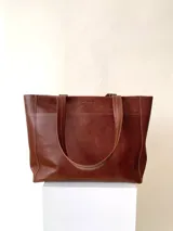 Cartera tipo tote de cuero graneado color negro, con textura uniforme y suave al tacto. Tiene manijas de 55 cm, bolsillo frontal con broche imantado, bolsillo trasero con cierre metálico, cierre principal metálico, forro interior y tres bolsillos internos (uno con cierre, uno plaqué y uno tipo sobre con solapa y botón).