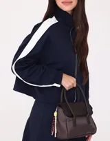 Cartera tipo city color beige, de tela deportiva, con doble asa corta, correa regulable desmontable, solapa con broche a presión y charm colgante.