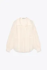 Camisa color crudo confeccionada en hilatura con mezcla de ramio, de corte holgado, con cuello solapa y escote pico. Tiene mangas largas con puños y detalle de tejido con pliegues.