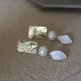 Aros colgantes hechos a mano en bronce, con pieza superior rectangular texturada color dorado, perla redonda color crema y pieza inferior en forma de hoja color blanco nacarado.