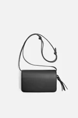 Bandolera negra con correa fina ajustable, cierre con solapa imantada, bolsillo interno y charm colgante.