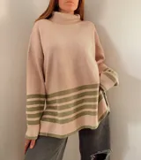 Sweater beige de tejido grueso con cuello alto, corte oversize y tajo a los lados. Tiene franjas horizontales verdes en la parte inferior.