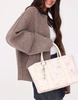 Cartera tipo tote color blanco crudo, con doble asa corta y correa larga regulable y desmontable. Incluye charm colgante con forma de corazón y cierre superior.