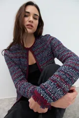 Chaqueta corta de tejido bouclé o tweed, sin cuello ni cierre frontal, con ribetes en color bordó. El tejido es una mezcla de hilos en tonos azul marino, rojo/bordó y blanco, creando un efecto jaspeado. Los puños tienen un detalle de flecos en color bordó.