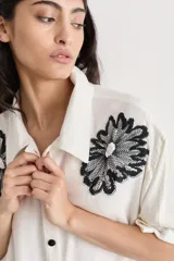 Camisa color crudo de corte amplio con bordados florales negros en la parte superior. Tiene cuello camisero, mangas cortas y cierre frontal con botones negros.