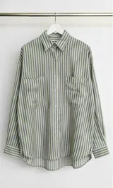 Camisa oversize a rayas verticales color verde y lila, con cuello clásico, cierre frontal con botones, mangas largas con puños abotonados y dos bolsillos de parche en el pecho.