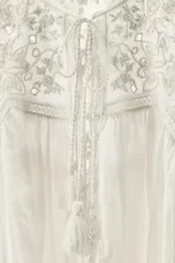 Blusa sin mangas color blanco con cuello alto, bordados y puntillas combinadas a tono, y apliques de abalorios. Cierre frontal con abertura, lazada y botón.