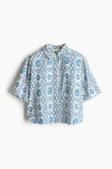 Camisa de corte holgado con estampado de flores azules sobre fondo blanco, cuello clásico, botones frontales y mangas cortas. Confeccionada en sarga con canesú y pliegue en la espalda, hombros caídos y bajo redondeado con aberturas laterales.