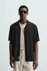 Camisa de calce relajado confeccionada en tejido fluido de mezcla de viscosa, color blanco. Presenta cuello bowling, manga corta, aberturas laterales en el bajo y cierre frontal con botones al tono.