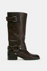 Bota de caña alta de cuero color marrón oscuro, estilo motociclista, con taco bajo y punta redonda. Presenta una tira con hebilla ajustable en la parte superior de la caña y una tira desmontable con argolla metálica y detalle de rosa repujada en el tobillo.