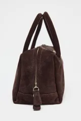Bolso formato bowling color marrón chocolate, confeccionado en serraje. Tiene doble asa de mano y correa bandolera extraíble. Incluye bolsillo interior con cremallera y cierre principal con doble cremallera con tirador.