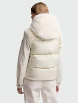 Chaleco Moncler modelo Arve en color blanco crema, con frente y capucha de pelo sintético. Presenta un diseño acolchado con cierre frontal de botones a presión negros, capucha ajustable con cordones y el logotipo de la marca bordado en el pecho.