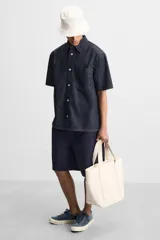 Bolso tipo shopper color crema, fabricado en tejido de algodón. Tiene doble asa corta, compartimento principal con cierre de cremallera, bolsillo interior con cremallera y múltiples bolsillos exteriores.