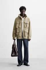Parka color beige de tejido técnico ligero, corte relaxed fit, cuello alto con capucha plegable, manga larga con puños ajustables con botón a presión, cierre frontal con cremallera, cintura ajustable con elástico interno y múltiples bolsillos frontales de parche con solapa.