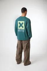 Remera de manga larga color verde azulado con cuello redondo. Presenta un pequeño logo de 'blue banana' en el pecho izquierdo, un cuadrado amarillo con una 'X' verde en el pecho derecho y texto en japonés verticalmente en la manga derecha.