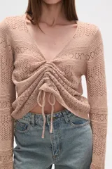 Sweater de poliéster color beige, con cuello en V, manga larga y frunces ajustables en el frente. Presenta un diseño de tejido calado.