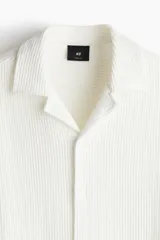 Camisa de manga corta color blanco, con tejido acanalado, cuello resort, tapeta lisa, canesú en la espalda y bajo recto con aberturas laterales.