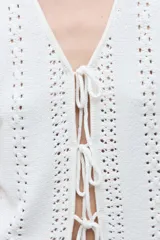 Chaleco sin mangas color blanco crudo, tejido a crochet o punto abierto, con escote en V y cierre frontal mediante lazos cruzados.
