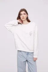 Buzo blanco de corte holgado con cuello redondo y mangas largas. Presenta un pequeño estampado de una flor en el centro del pecho.