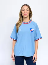 Remera blanca de corte clásico con cuello redondo y mangas cortas con puños tricolor (azul, blanco y rojo). Presenta el logo de Umbro bordado en el pecho izquierdo y el escudo del Club Nacional de Football bordado en el pecho derecho.